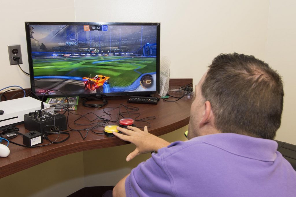 A patient in a power chair seen from behind, playing a racing game on the Xbox One using buddy button adaptive controllers