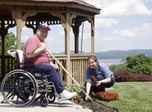 Man in wheelchair using adaptive gardening tool with long pole to reach flower garden bed. Women kneeling down opposite him. In front of a gazebo in Helen Hayes Hospital's MacArthur Park, overlooking the Hudson River.