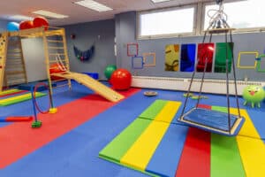 Photo of Helen Hayes Hospital pediatric rehabilitation suite, showcasing the suite's wooden sensory gym, with climbing elements and a slide, and a therapy swing hung from the ceiling. There are a variety of colorful pieces of therapy equipment in the room, including blue, red, and green balls and colorful wall hangings.