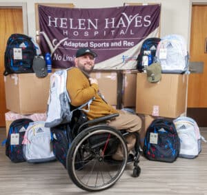 Helen Hayes Hospital's Adapted Sports and Recreation Coordinator, Peter Gagliardo, is seated in his wheelchair with backpacks, water bottles, and hats donated by Adidas surrounding him. He has a backpack slung over his shoulder and the Helen Hayes Hospital Adapted Sports & Recreation Program banner hanging behind him which features the tagline "no limitation." 