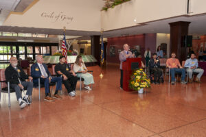 New York State Commissioner of Health, Dr. James McDonald, provides congratulatory remarks at the 125th annual Helen Hayes Hospital Honors Assembly. Dr. McDonald stands at a podium, flanked by the 8 honorees in chairs on either side of the podium. 