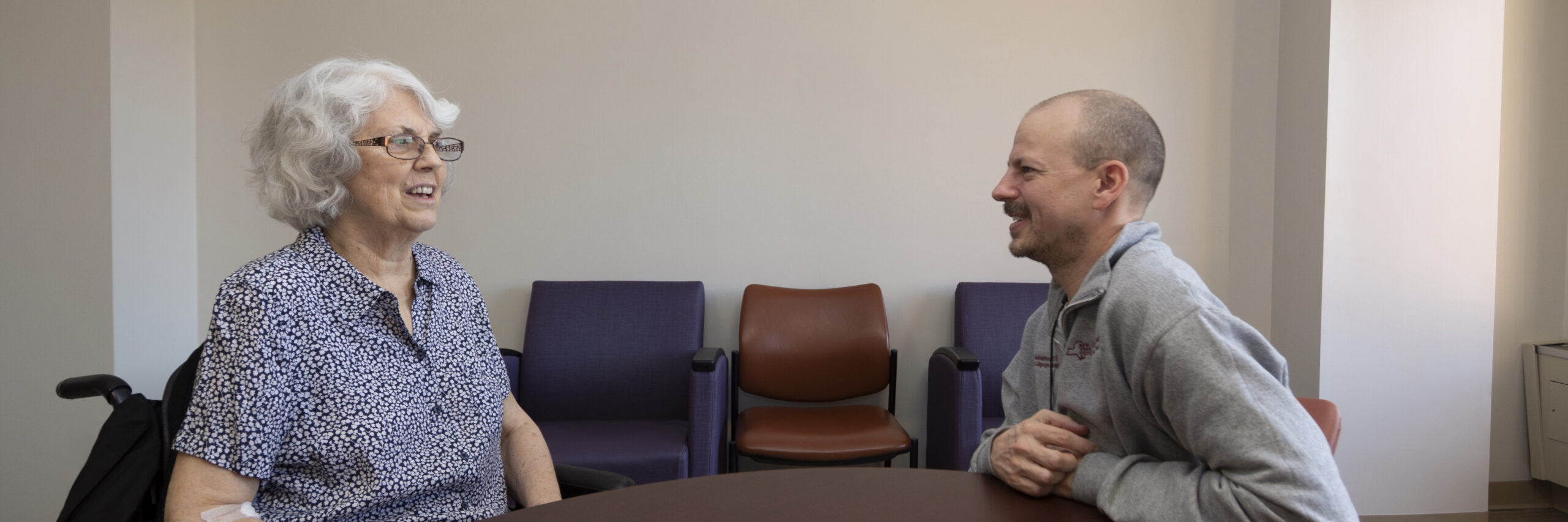 A female patient in a wheelchair sits across from a male speech therapist at a wood table. Both are smiling as they converse.