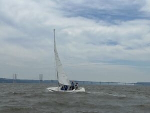 A sailboat from the Nyack Boat Club's Sonar Fleet 23 sailing on the Hudson River with the Mario Cuomo Bridge in the background and a blue and cloudy sky above. The boast is heading left and heeling to the left side.
