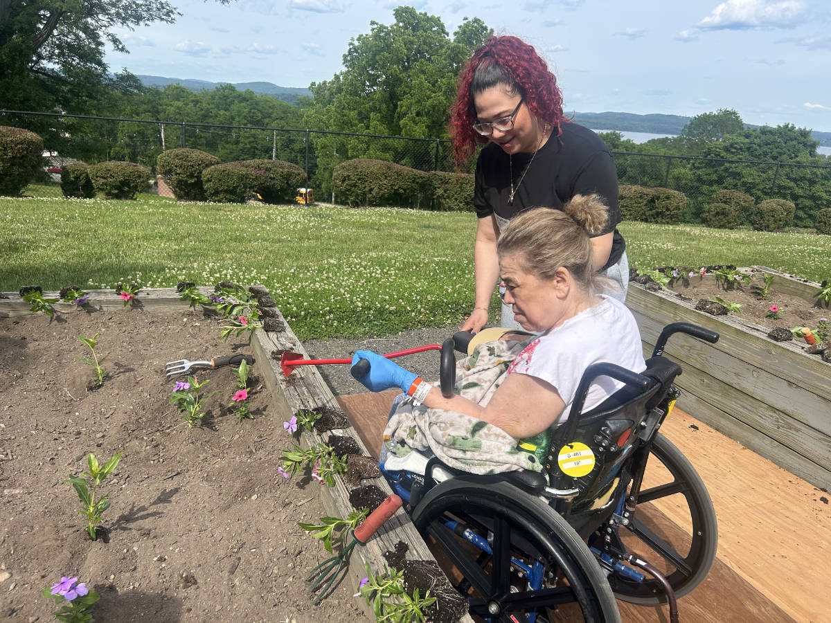 A woman in a wheelchair sits to the right of a raised gardening bed. In her hand is a long handled adaptive gardening trowel. Her daughter stands behind her, smiling and ready to support her. The garden box is filled with colorful vinca flowers. It is a sunny day and the Hudson River is visible in the distance.