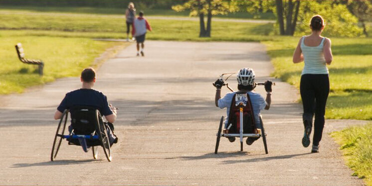 Two men riding handcycles on a path outside with a woman running alongside them