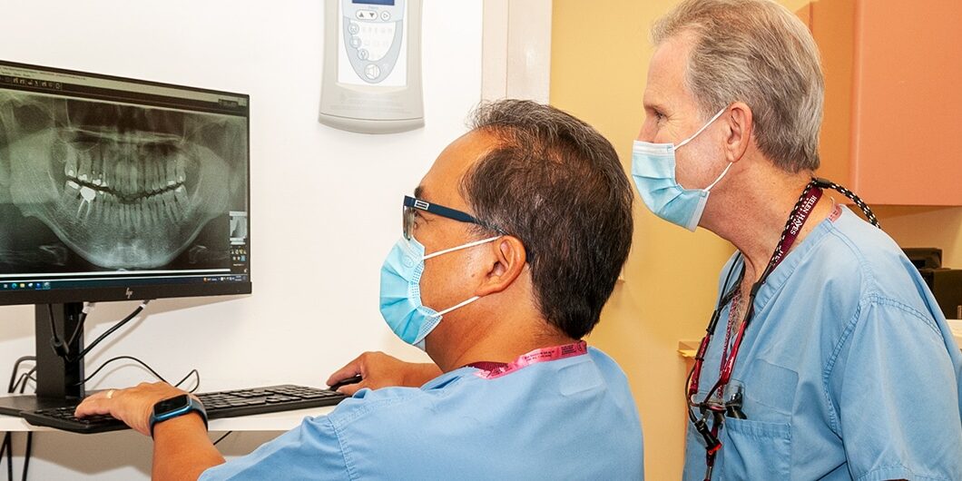 Dr. Nestor Santos sitting and Dr. Marc Michalowicz standing and reviewing a tooth X-ray on a computer screen