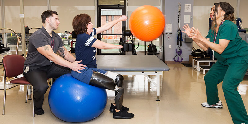 Female amputee sitting on a balance ball while throwing a ball for the physical therapist to catch